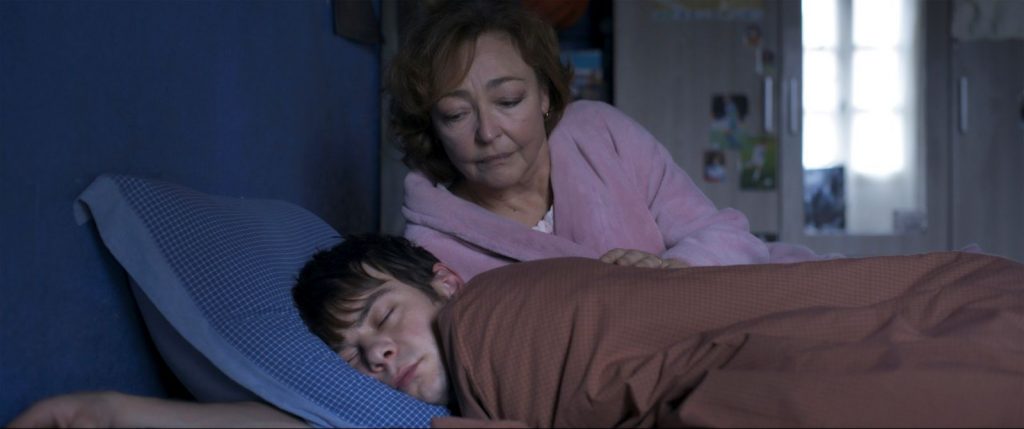 A young man sleeps in bed while an older woman peers over him.