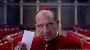Ralph Fiennes in the film Conclave. He is wearing a Cardinal's red robe and cap, and holding a white ballot with a very serious look in his eyes.