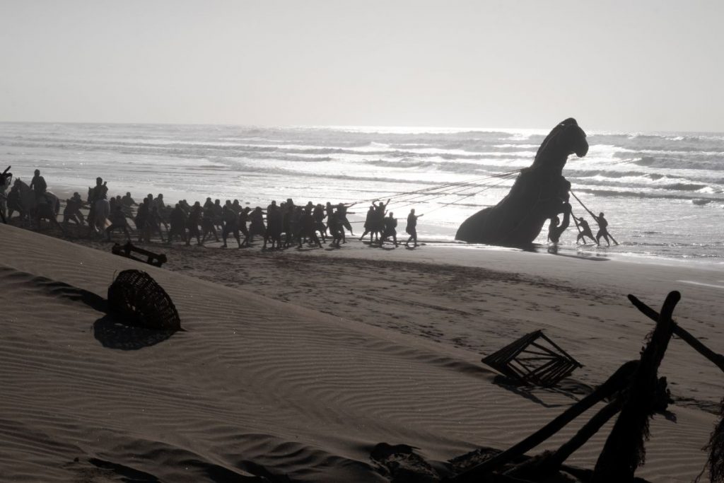 A wide shot shows an army of soldiers in ancient times on a beach with a Trojan horse at the water's edge.