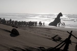 A wide shot shows an army of soldiers in ancient times on a beach with a Trojan horse at the water's edge.